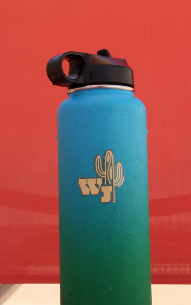 WJ Saguaro Sticker in teal with yellow lettering in addition to the wildjoy lettered on the arm of the saguaro. It is on a teal waterbottle with a black lid. It's resting on a red chair.